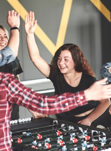A diverse group of cheerful coworkers celebrating together while playing a game of foosball in a modern office environment.