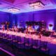 Elegant banquet hall set up for a corporate awards event with purple lighting, decorated tables, floral centerpieces, and candles.