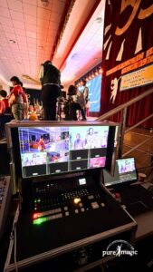 A professional vision mixer and multi-view monitor display live camera feeds during an on-stage performance, demonstrating the AV equipment necessary for successful hybrid event production.